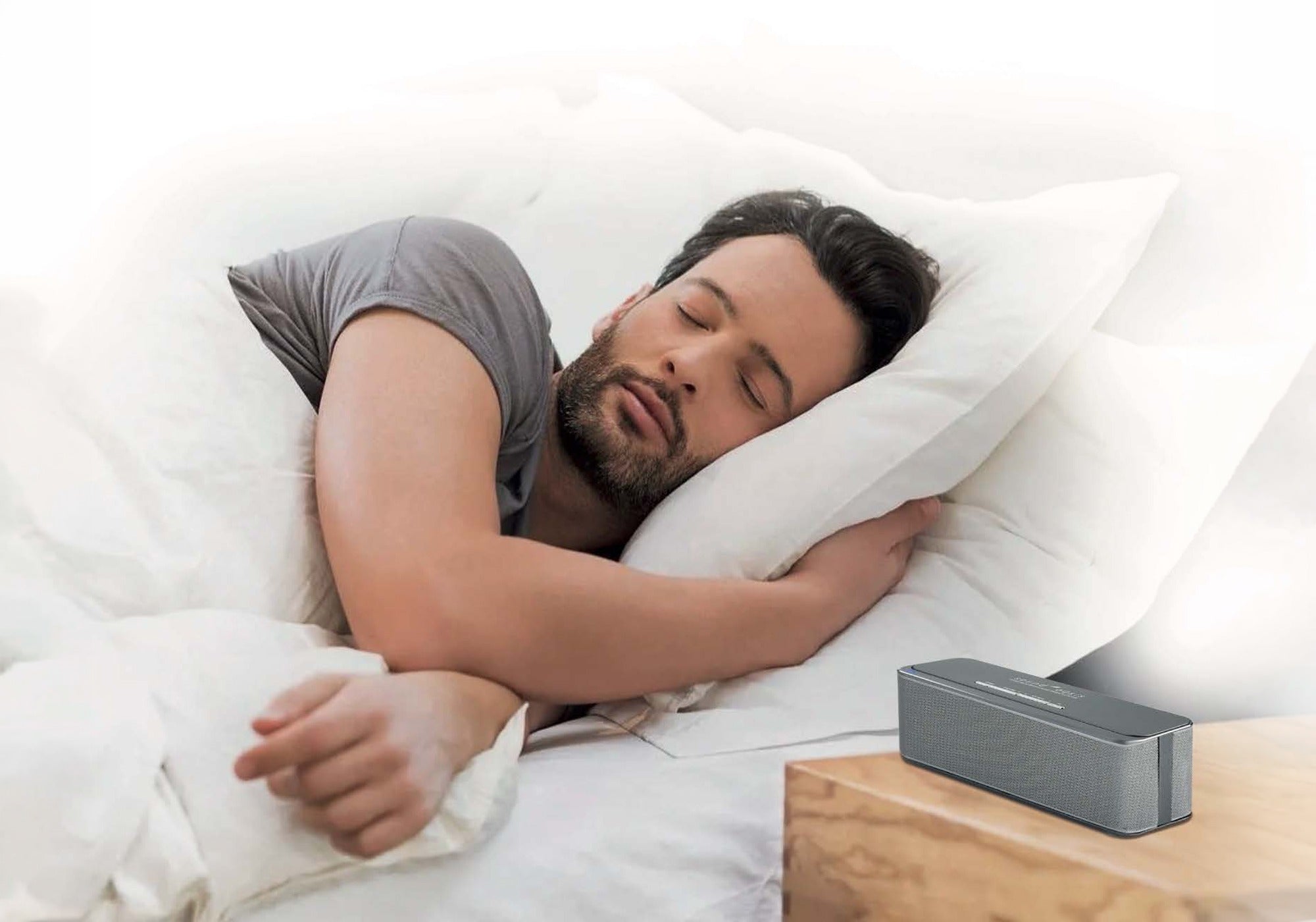 A sleeping man rests on a white pillow and bed, with a gray rectangular electronic device on a wooden bedside table nearby.