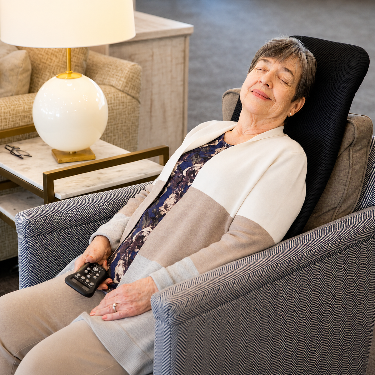 An older woman reclines comfortably in a gray textured armchair, holding a black remote control. She is wearing a beige and white color-block cardigan over a navy floral top.