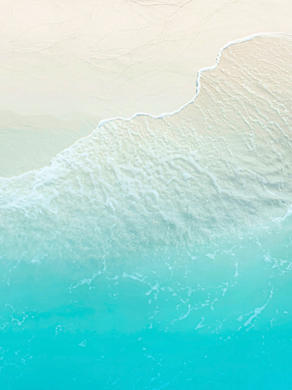 Abstract aerial view of turquoise ocean water meeting pale sand, with white surf and a soft diagonal shoreline.